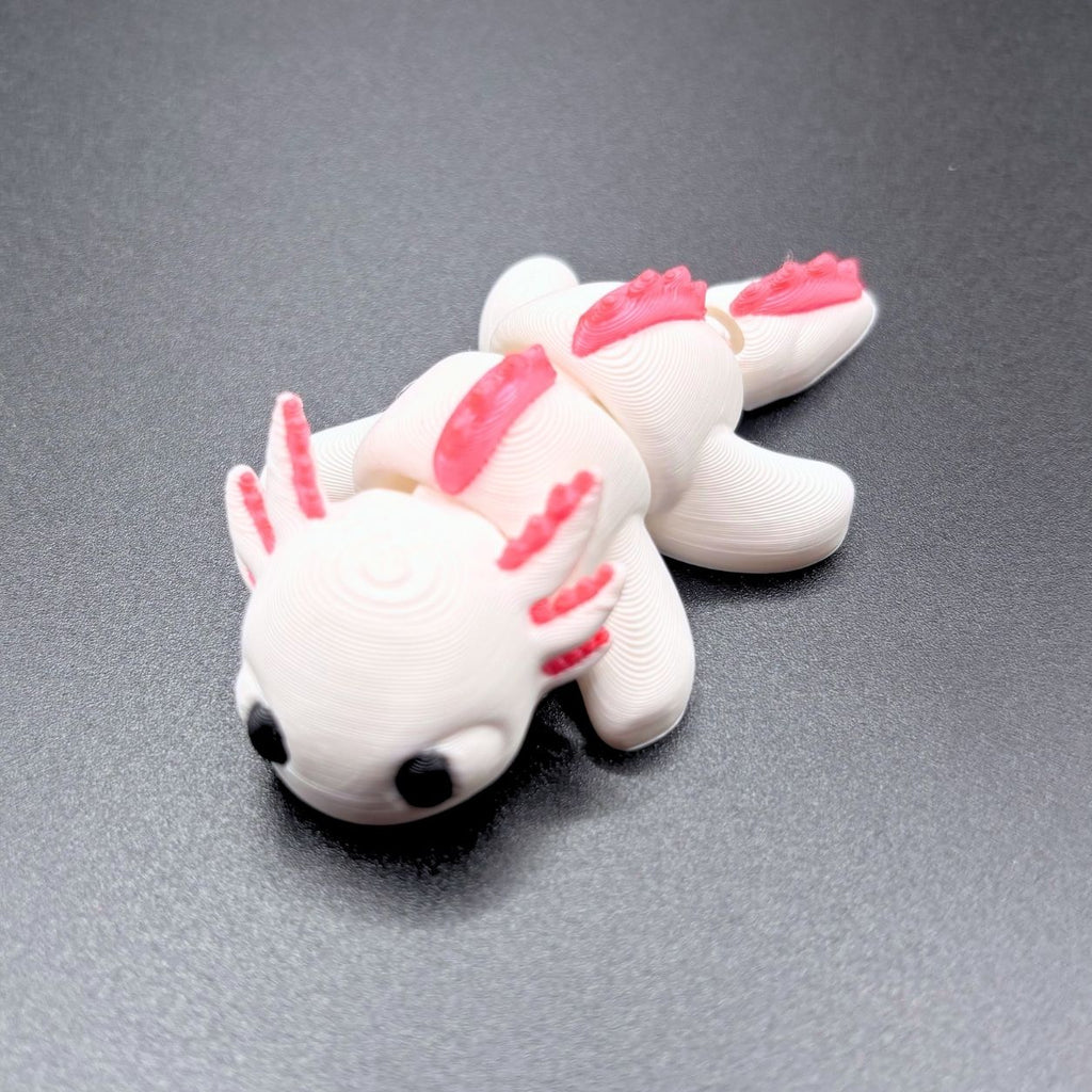 A small white 3D-printed axolotl figurine with bright pink gills and fins, resting on a dark gray textured surface.