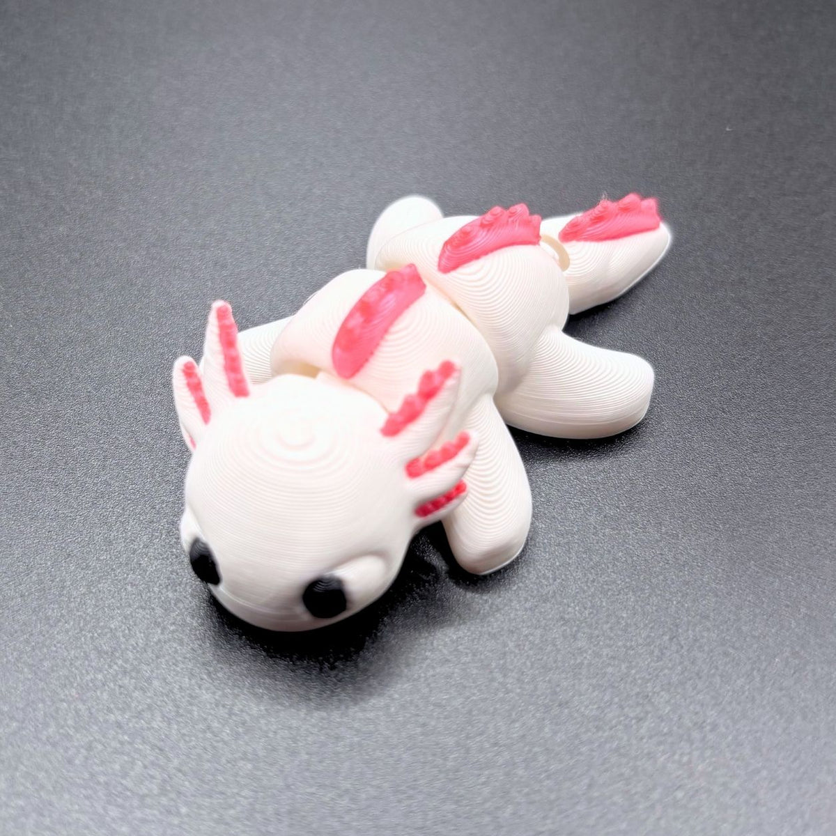 A small white 3D-printed axolotl figurine with bright pink gills and fins, resting on a dark gray textured surface.