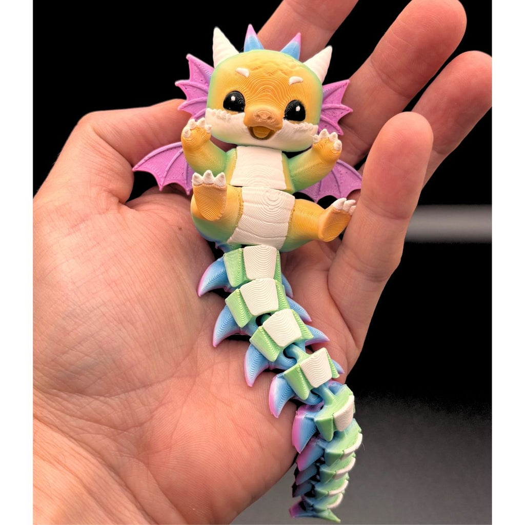 A small colorful dragon 3D being held in a person's hand. The dragon has yellow, green, white, pink, and blue details, with pink wings and a segmented tail, photographed against a dark background.